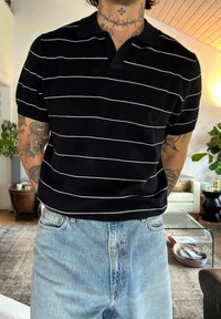 Hombre con tatuajes en el brazo y el cuello, vistiendo una camiseta polo negra con rayas blancas y vaqueros de color azul claro, de pie en una sala de estar.