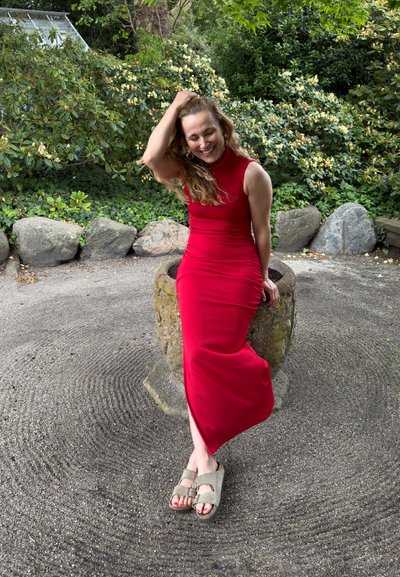 Vestido ajustado rojo con cuello alto, acentuando la silueta. Llevado con sandalias beige, sentada sobre una piedra con un suelo texturizado. Fondo de follaje verde.