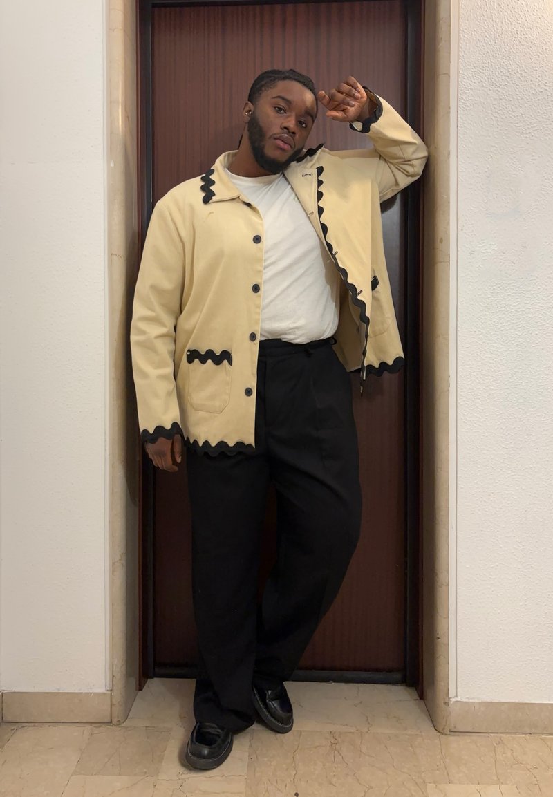 Uomo appoggiato a un telaio di porta in legno, indossando una giacca beige con rifiniture nere, una camicia bianca, pantaloni neri e scarpe nere, posando con un braccio alzato.