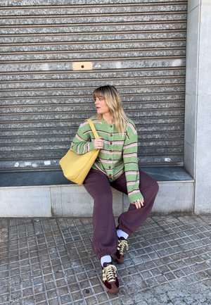 Joven mujer con suéter verde a rayas y pantalones morados, sentada en un borde con un bolso amarillo, mirando hacia su izquierda frente a una persiana metálica.