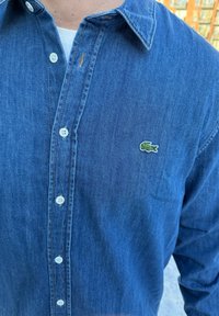 Sötétkék farmering hosszú ujjal, hegyes gallérral és fehér gombokkal. A bal mellrészen kis zöld Lacoste logó található.