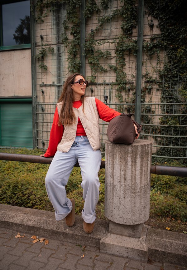 Mujer con gafas de sol, suéter rojo y chaleco beige sentada en una barandilla de metal al aire libre, apoyando la mano en un poste de cemento con bolso marrón.