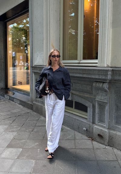 Camisa de manga larga a rayas negras, pantalones de lino blancos, sandalias negras, sosteniendo un bolso texturizado con adornos decorativos, de pie en la acera.