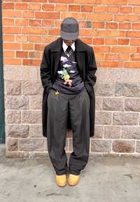 Must pikk mantel, must nokamüts, graafilise musta t-särgiga, millel on värvikas disain, hallid liibuva lõikega püksid ja beežid klotskingad, seismas tellisseina ees.