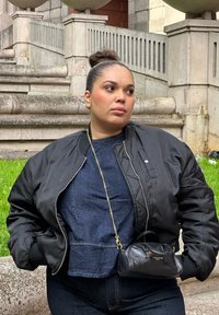 Crna bomber jakna preko tamnoplave traper košulje, uparena s tamnim trapericama. Mala crna torbica sa zlatnim lancem, postavljena na pozadini zelene trave.