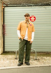 Ein orange gestreiftes Kurzarmhemd über einem weißen Langarmhemd, olivgrüne weit geschnittene Hose, schwarze Schuhe, mit einer Mütze und einem Schal als Accessoire.