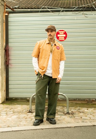 Camisa de manga corta a rayas naranjas sobre una camisa de manga larga blanca, pantalones anchos verde oliva, zapatos negros, con una gorra y una bufanda como accesorios.