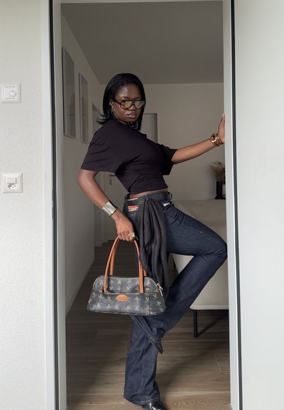 Mujer con camiseta negra y jeans oscuros posando en un marco de puerta, sujetando un bolso estampado con asas marrones, y llevando gafas de sol y joyas.