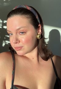 Top nero con spalline sottili, orecchini circolari dorati e una fascia per capelli di tessuto scozzese. Pelle liscia, trucco leggero, onde morbide nei capelli.