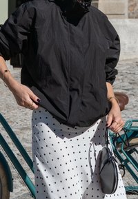 Čierna ľahká bunda s vysokým golierom, v kombinácii s bielou sukňou so zelenými bodkami. Drží malú čiernu kabelku.