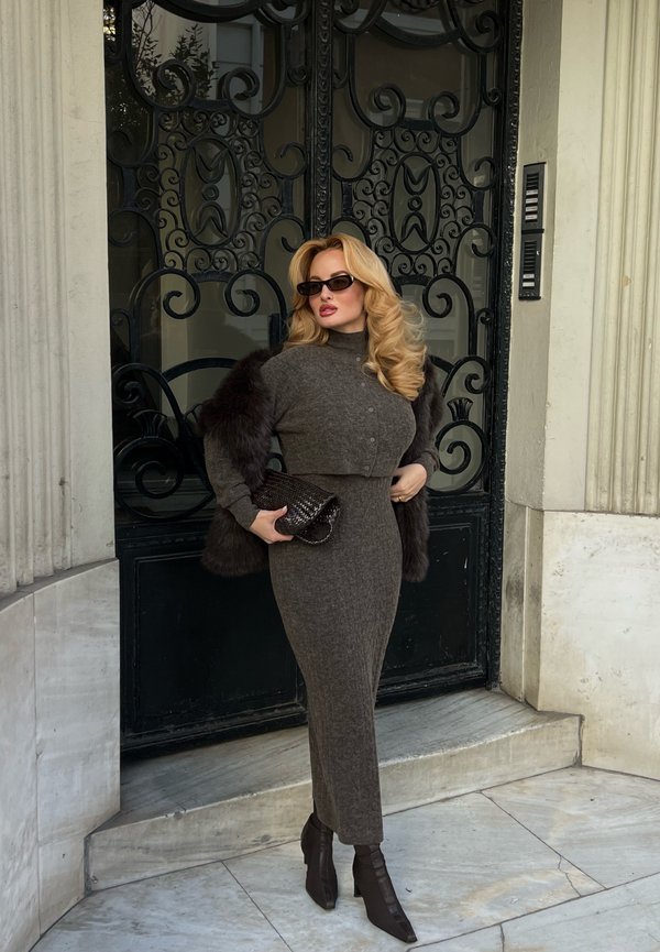 Femme aux cheveux blonds bouclés portant une robe grise moulante et un stole en fourrure, tenant une pochette noire, debout devant une porte en métal noir ornée.