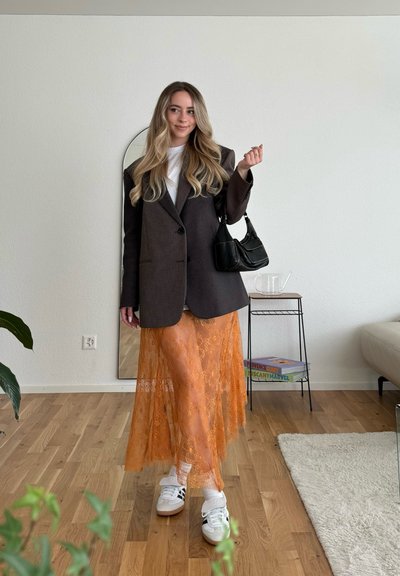 Blazer marrón con patrón, camisa blanca, falda de encaje naranja y zapatillas blancas. Bolso negro con asa curva. Entorno minimalista con plantas.