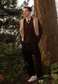 Νέος άνδρας στηρίζεται σε μεγάλο δέντρο στο δάσος, φοράει σκούρο πουλόβερ, μπεζ πουλόβερ ριγμένο στους ώμους, μαύρο παντελόνι και λευκά αθλητικά παπούτσια.