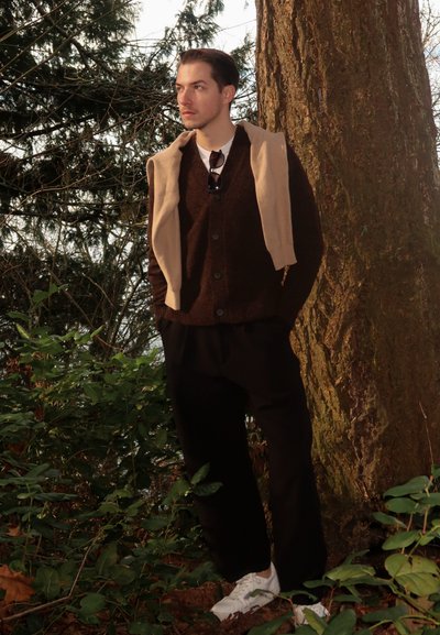Joven apoyado en un gran árbol en el bosque, vistiendo un suéter oscuro, un suéter beige sobre los hombros, pantalones negros y zapatillas blancas.