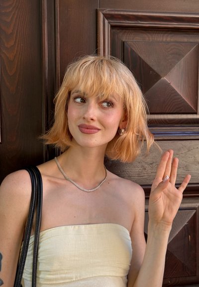 Top crema sin tirantes, collar de cadena plateada y bolso negro. Pelo rubio corto y ondulado. De pie frente a una puerta de madera con patrones geométricos.