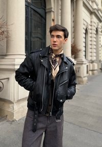 Črna usnjena motoristična jakna s srebrnimi zadrgami, v kombinaciji s svetlo rjavo srajco in vzorčastim šalom; model stoji ob arhitekturni kulisi.