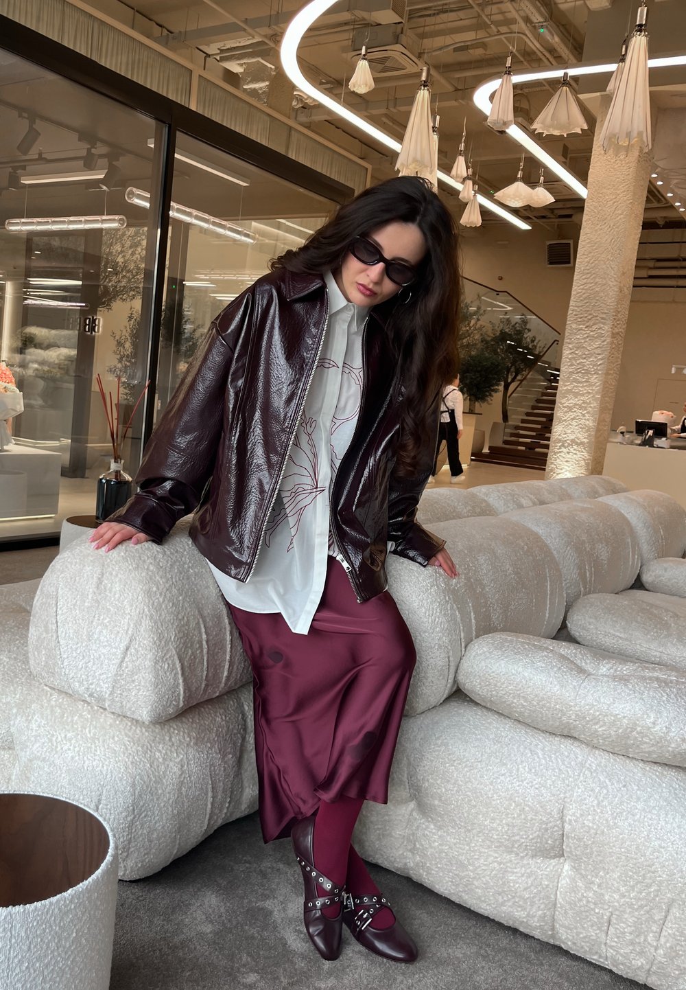 Bordeaux, glanzende leren jas over een wit shirt met bloemenpatroon, gecombineerd met een satijnen bordeaux rok en bijpassende schoenen met hardware-accenten.