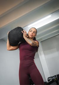 Black sandbag, worn on the shoulder, with a smooth texture. The athlete is wearing a fitted maroon top and high-waisted maroon leggings with side stripes.