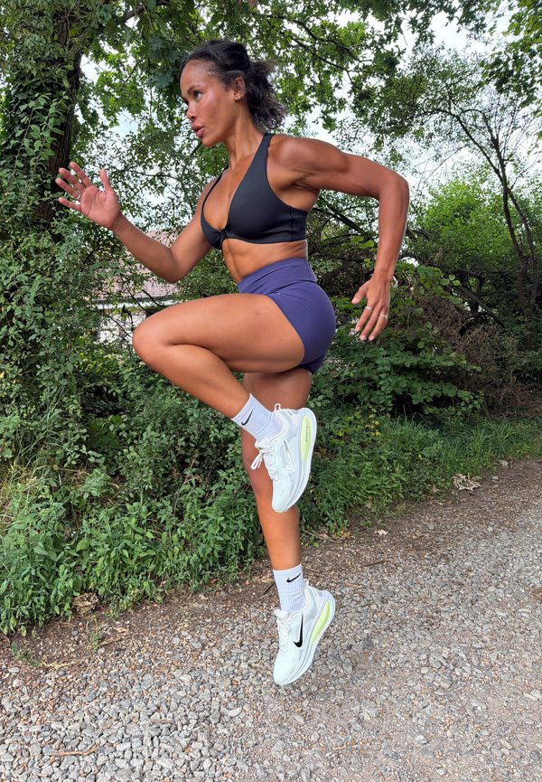 Donna atletica in abbigliamento sportivo che esegue un esercizio di ginocchio alto su un sentiero ghiaioso circondato da alberi e cespugli verdi.