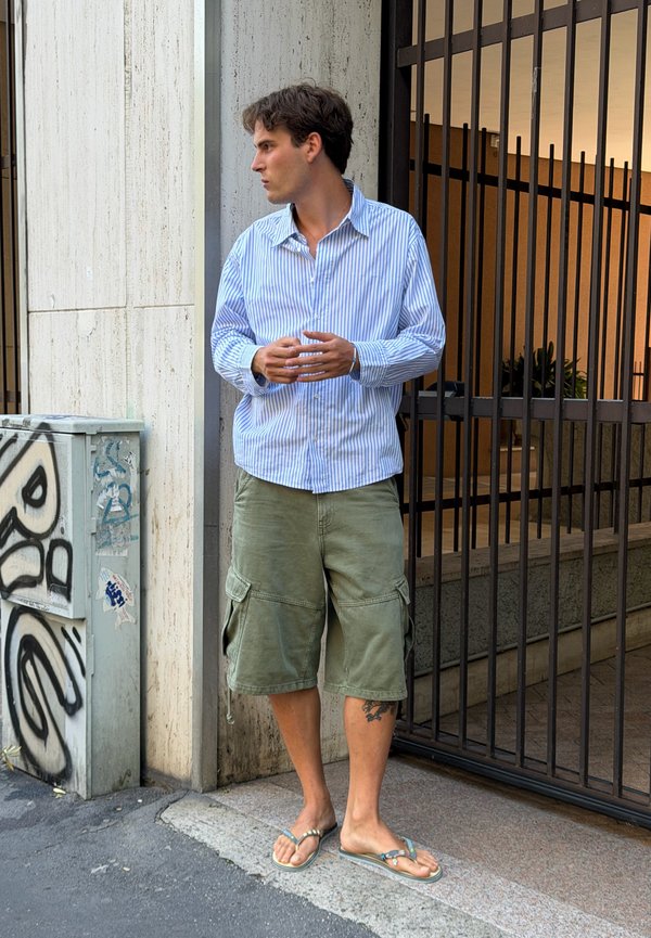 Giovane uomo con camicia a righe, pantaloncini cargo verdi e infradito, in piedi sul marciapiede vicino a un cancello e a una cassetta degli impianti coperta di graffiti.