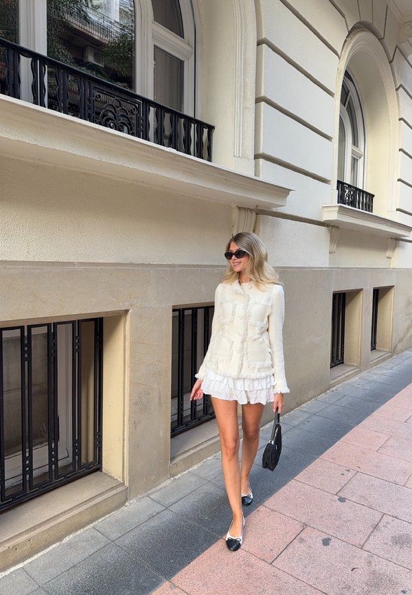 Witte textuurtrui met franjes, over een gerimpelde witte rok. Een zwarte handtas en zwart-witte platte schoenen maken de outfit compleet.