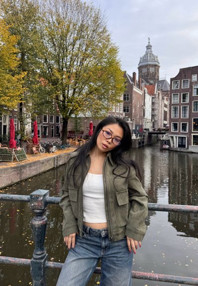 Mujer joven con gafas y chaqueta verde apoyada en la barandilla de un puente sobre un canal, con árboles de otoño y edificios históricos de fondo.