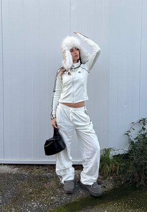 Mujer con chándal blanco y gorro de piel posa al aire libre, sosteniendo una pequeña bolsa negra, de pie sobre grava cerca de una pared de metal y plantas.