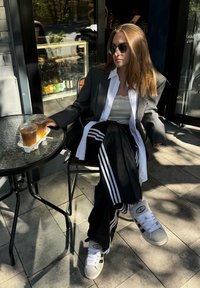 Calças de pista em preto e branco com três riscas, um blazer oversized cinza, uma camisola de renda branca e ténis cinzentos, sentado a uma mesa de vidro.