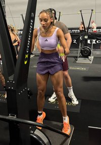 Top deportivo corto morado y pantalones cortos oscuros, con zapatillas deportivas naranjas. Se ve una máquina de ejercicio negra en un gimnasio, enfocada en la actividad física.