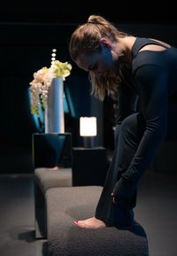 Fekete sportöltözetbe öltözött nő a lábát egy padra nyújtja egy gyér világítású szobában, a háttérben egy virágcsokor és egy lámpa látható.