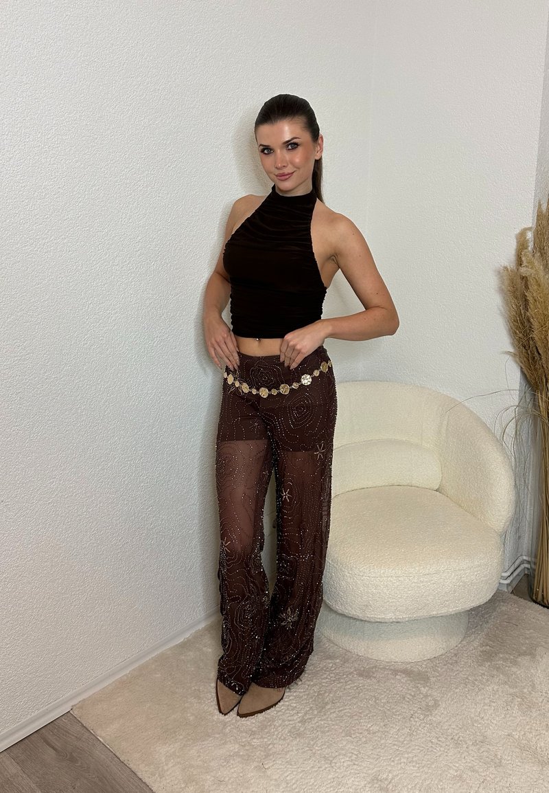 Femme debout à l'intérieur près d'une chaise blanche texturée, portant un haut noir sans manches, un pantalon sombre transparent avec des motifs circulaires, et des chaussures pointues beiges.