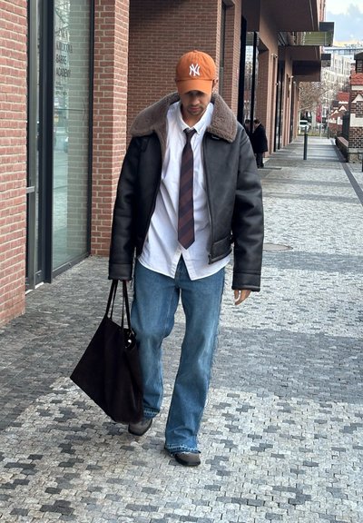 Chaqueta de cuero negra con cuello de piel sintética, camisa blanca, corbata oscura, jeans de mezclilla azul claro y gorra naranja, llevando una bolsa tote negra mientras camina por un pavimento de piedra.