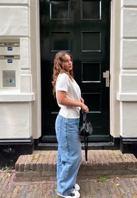 Hvit kortermet topp kombinert med lyseblå vidde jeans. En svart veske holdes i hånden. Bakgrunnen har en mørkegrønn dør og murtrapp.
