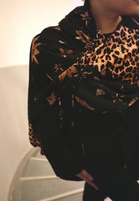 Personne portant un manteau noir et une écharpe noire décorée de motifs floraux et léopard marron, debout près d'un escalier courbe.