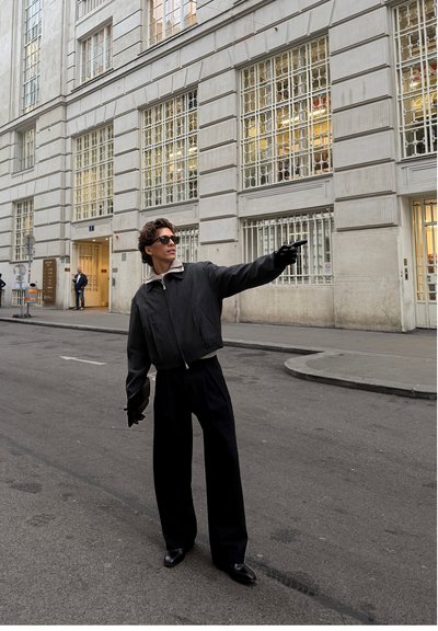 Cazadora bomber negra recortada con cuello gris, pantalones negros de talle alto y gafas de sol, de pie en una calle frente a una fachada de edificio moderno.