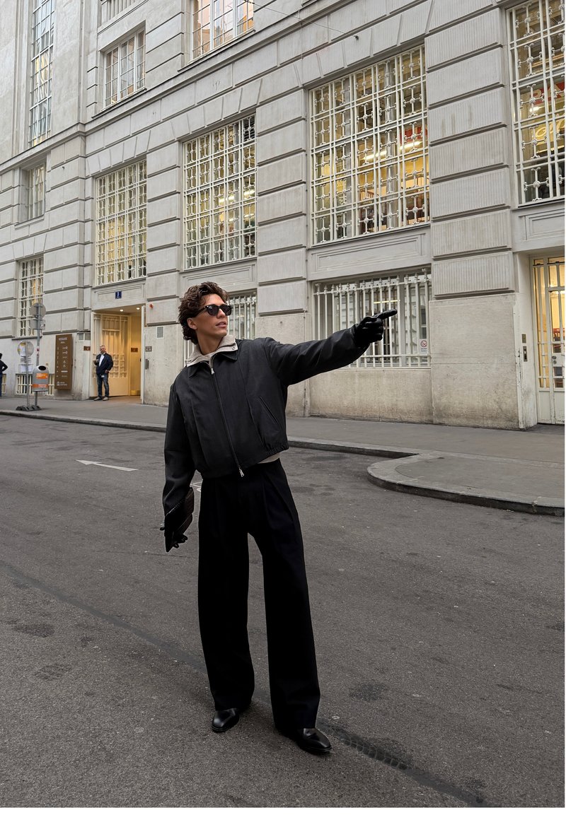 Schwarze, bauchfreie Bomberjacke mit grauem Kragen, hoch taillierte schwarze Hose und Sonnenbrille, stehend auf einer Straße mit moderner Gebäudefassade.