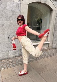 Top cropped vermelho com bolinhas brancas, calças capri bege, tamancos vermelhos com plataforma e detalhes brancos e laço; segurando uma pequena mala vermelha contra uma parede de pedra.