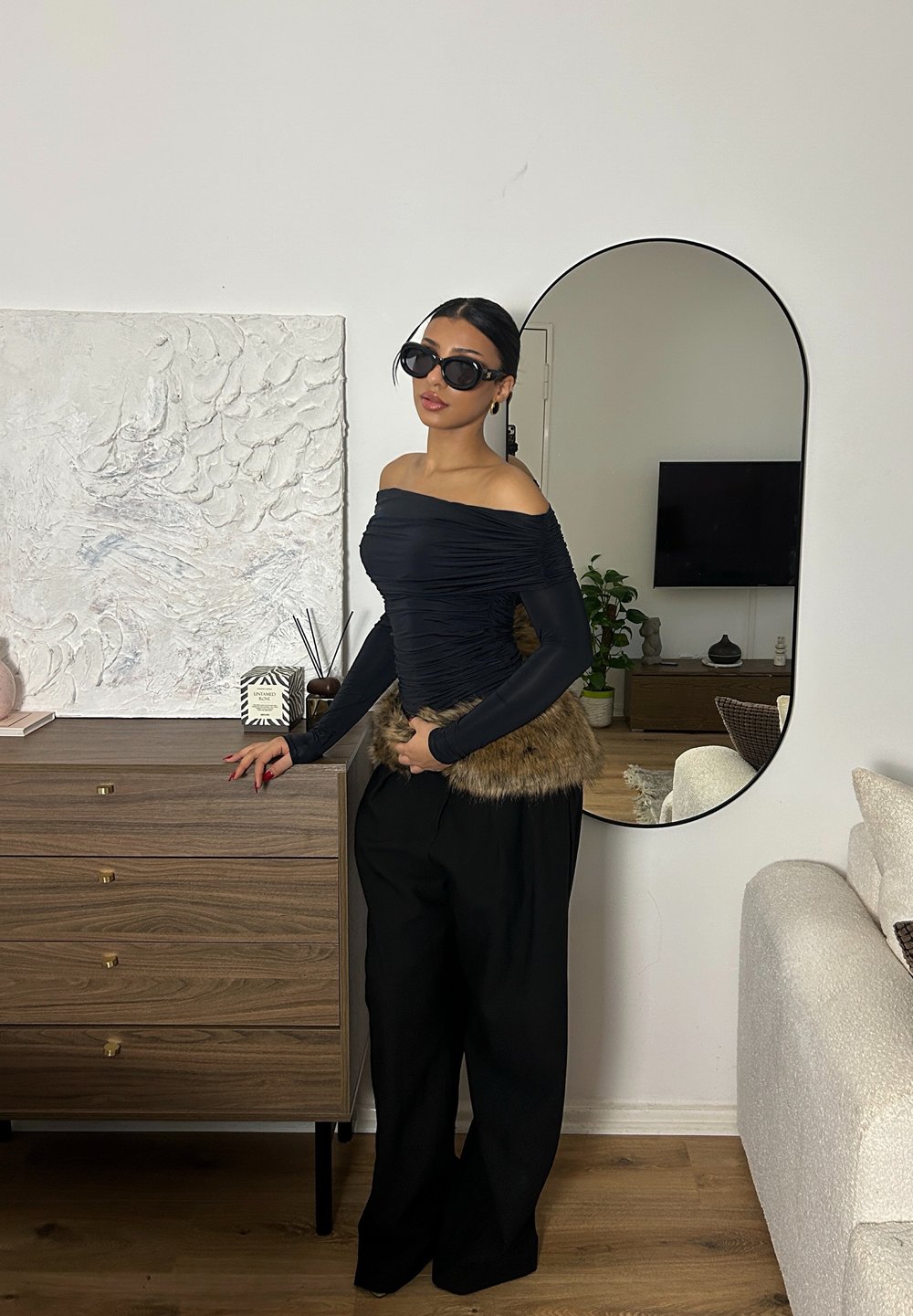 Sort off-shoulder top med teksturerede ærmer, kombineret med brede sorte bukser og en brun pelsdetalje. Tilbehør inkluderer store sorte solbriller.