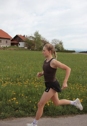 Una joven corre por un sendero, vistiendo una camiseta sin mangas marrón y pantalones cortos negros. Lleva zapatillas deportivas blancas, con un paisaje natural de fondo.