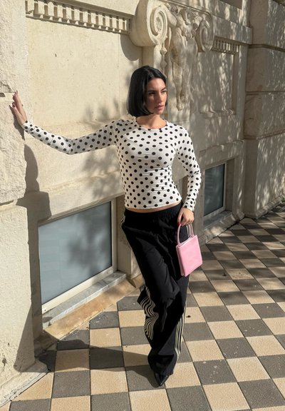 Mujer con cabello corto y negro, vestida con una blusa blanca con lunares y pantalones negros, sosteniendo una bolsa rosa, apoyada en un muro de piedra bajo la luz del sol.