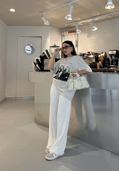 Camiseta gris de gran tamaño con un gráfico, pantalones de pierna ancha blancos texturizados, zapatos gris claro con rayas, sosteniendo un bolso transparente y una bebida verde.