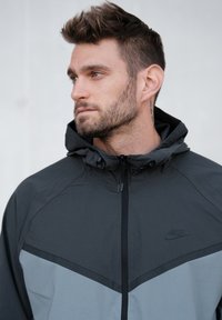 Kurtka Nike w ciemnoszarym i jasnoszarym kolorze, wyposażona w kaptur, zapięcie na zamek oraz logo na piersi. Materiał o fakturze z sportowymi liniami designu.