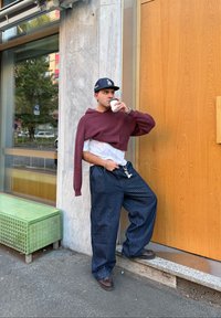 Bordový pletený svetr draped přes bílou košili, námořnické pruhované kalhoty s volnými nohavicemi, hnědé boty, držící šálek kávy, stojící proti dřevěné zdi.
