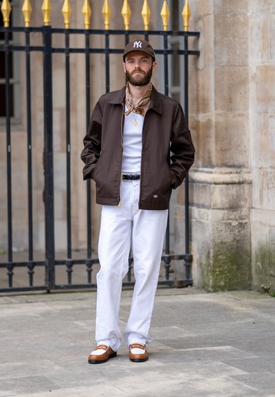 Chaqueta marrón, camiseta sin mangas blanca, pantalones blancos, zapatos marrones con acentos blancos. Accesorios incluyen una gorra y una bufanda. De pie frente a una valla.