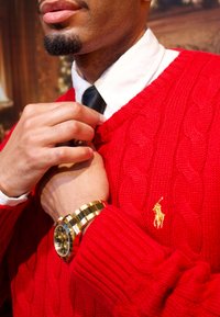 Pull-over rouge en maille torsadée avec un petit logo doré, porté sur une chemise blanche et une cravate noire. Une montre en or orne le poignet, complétant le look.