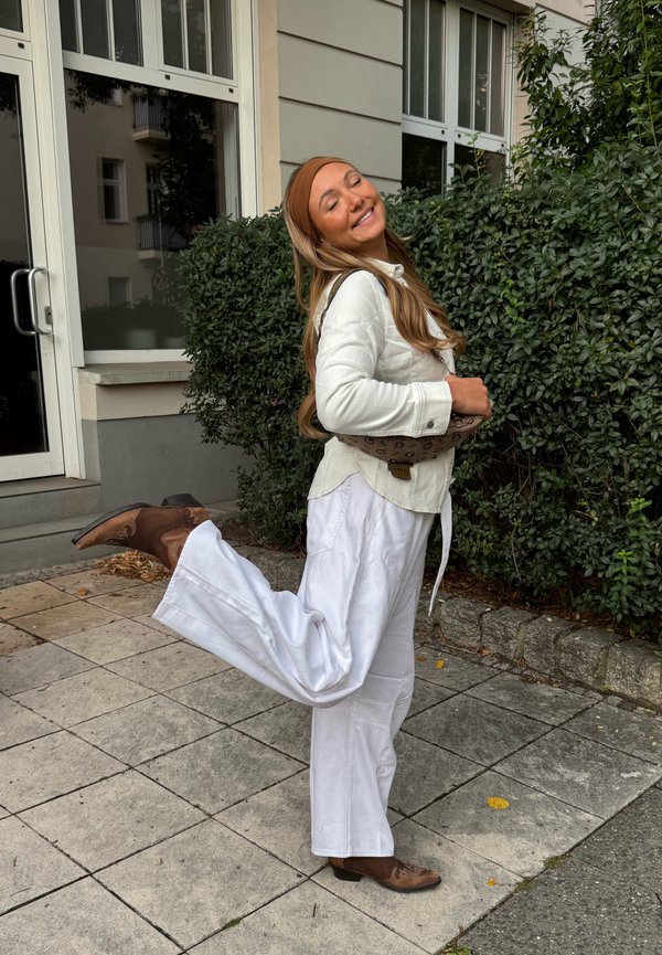 Femme souriante les yeux fermés, debout sur une jambe à l'extérieur près d'un bâtiment et des buissons, portant des vêtements blancs et des bottes marron.