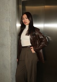 Γυναίκα με μακριά σκούρα μαλλιά φοράει λευκό ζιβάγκο, καφέ δερμάτινο μπουφάν και ψηλόμεσο χακί παντελόνι, στέκεται μπροστά σε μπετονένιο τοίχο και μεταλλική πόρτα.