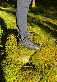 Svart sportsko med teksturert overflate og forsterket tå, plassert på et mosegrønt underlag, paret med mørke bukser.