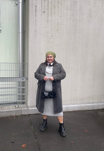 Abrigo gris a rayas, gorro de lana verde, vestido gris, bolso negro y botas de combate, enmarcados contra una pared texturizada y pavimento.