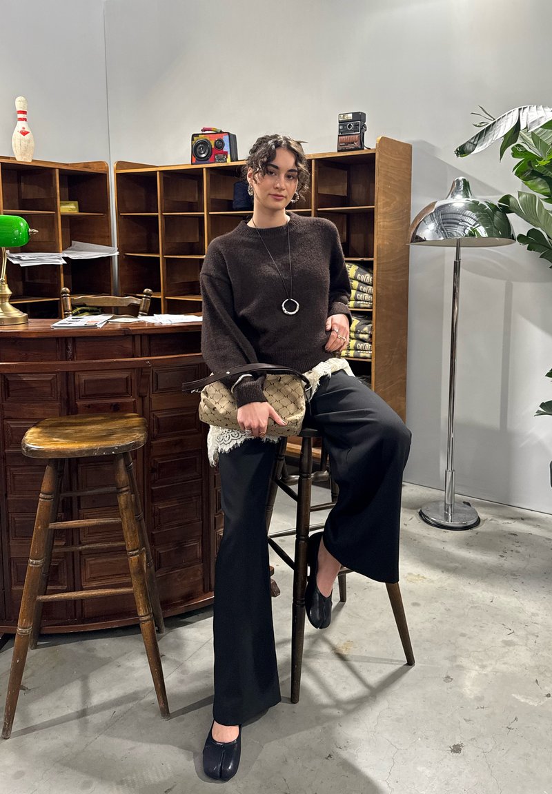 Cozy brown sweater, black wide-leg pants, holding a textured beige bag with lace trim. Setting features wood bar and vintage decor.